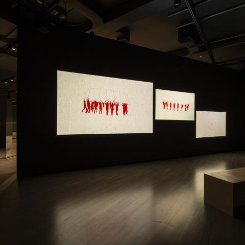 Mirrors connected by a long red string are in the background. There is a wall with three screens projected onto it. Each of the screens features red and white animations. There is one wooden bench in the foreground.