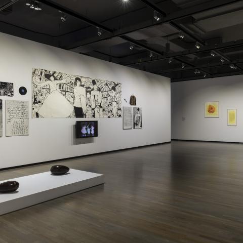 Two sculptures sit on a platform in the foreground of the image. A mixed media display is on the wall including, drawings, a record, an items of clothing and a TV screen. Five framed artworks hang on another wall in the background.