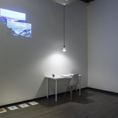 Four pieces of paper with earth are on the ground next to a white desk with a white chair. There is a binder on the desk and a single light bulb hangs above it on a black cord. Two images are projected onto the wall above the desk to the left.