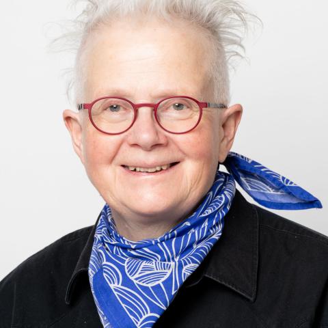 Rita McKeough smiles at the camera wearing red glasses and a blue scarf