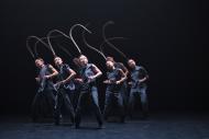 a group of 7 asian dancers on stage, wearing black clothes adn a long bird feather on their heads 