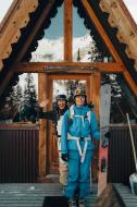 Two women with ski gears are standing in a front of a contemporary chalet door. The snowy high peaks, mountains and trees are reflecting into the windows 
