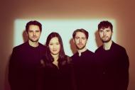 Four musicians dressed black, standing in front of a backlit wall