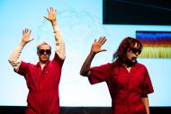 Two musicians in bright red coveralls perform in front of a blue backdrop