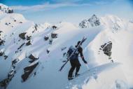 Alpine skier ascends a steep, snowy mountain ridge with tools and skis strapped to their backpack. 