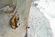 Man wearing yellow jacket climbing a big wall. Background shows a second man and glacier at the bottom. 