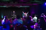 Audience seated at cabaret-style tables watches two performers on stage under colorful purple and green lighting during a live show.