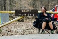 Woman wearing sport clothes and running shoes is seating in a camping chair in front of a trail head with an old wood sign indicating ''Do not block Gate''