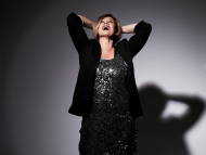 Woman wearing black suit and sparkly top pose with projector light. She is smiling looking up with her hands behind her back 