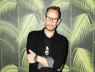 Portrait of a DJ standing confidently with arms crossed in front of a tropical palm leaf backdrop.