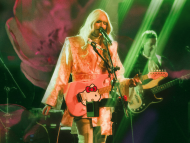 Performer with long platinum-blonde hair sings into a microphone while playing a pink electric guitar under green stage lights, with another musician performing in the background.