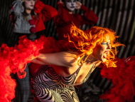 High-energy cabaret performance featuring a singer with vibrant red hair and dramatic red costume elements, surrounded by theatrical performers in an immersive stage setting.
