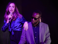 Singer holding a microphone stands beside a pianist wearing sunglasses under purple stage lighting during a live performance.