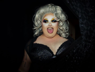 Performer wearing dramatic stage makeup, a platinum-blonde wig, and a black sequined outfit poses with their tongue out against a dark background.